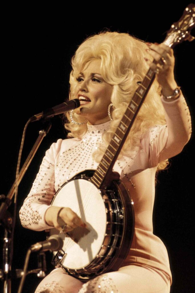 Dolly Parton, a white woman, sits on stage to perform her banjo and sing.