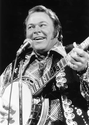 A black and white image of Roy Clark, a white man, as he sings and plays his banjo onstage.