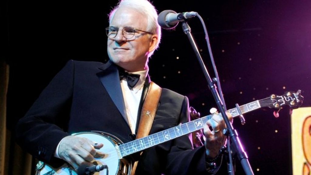 Steve Martin, an older white man, is on stage holding a banjo.