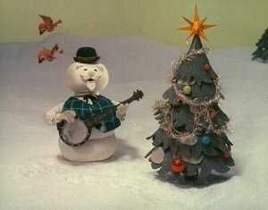 A stop motion snowman next to a Christmas tree holds his banjo and sings.