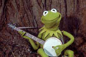 Kermit, a green frog puppet, sits against a tree holding a banjo.