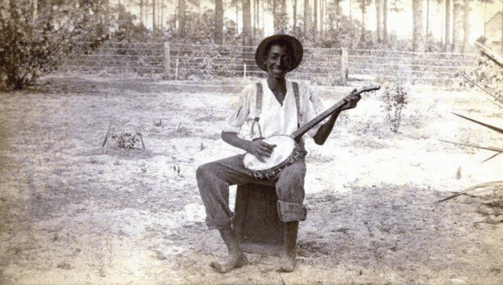 An old, black and white photograph of a black person sitting in the middle of the frame holding a banjo and smiling.