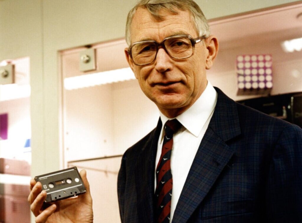 Lou Ottens poses for a camera, holding a cassette tape in his hand.