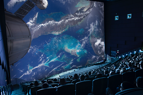 Large movie theatre with a very large and rounded screen with people in the audience. A satellite in space is projected on the screen.
